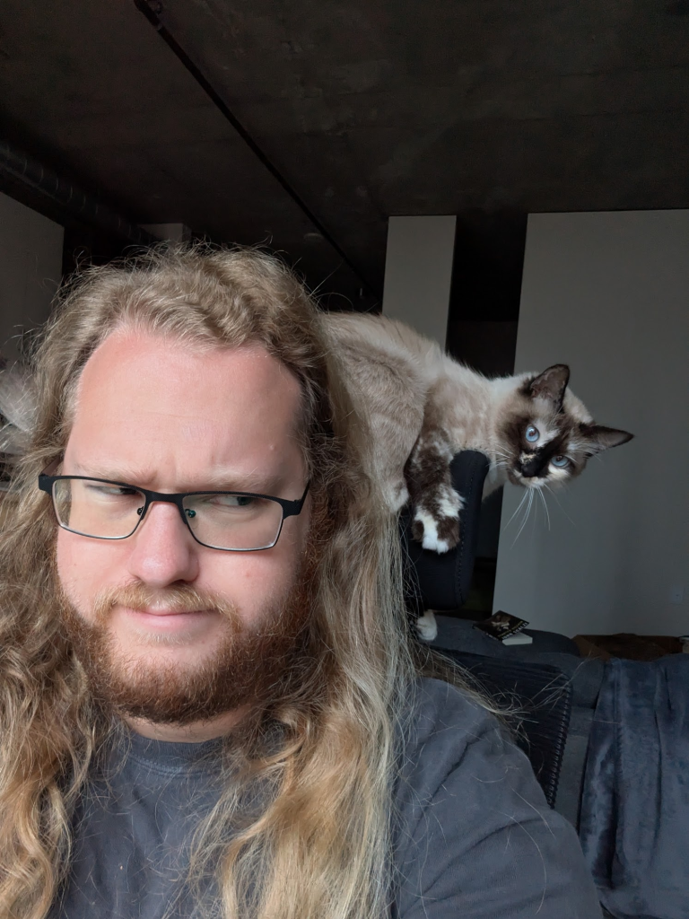 Ben sitting on a chair, with Marshmallow clinging to the headrest. Marshmallow is looking at the camera, while Ben is looking sideways at her in confusion.